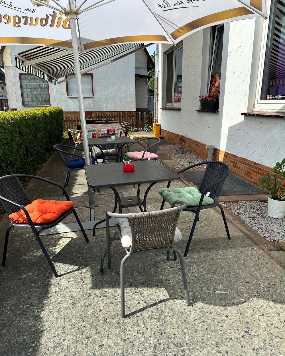 Sonniger Außenbereich von Pizzeria Aroma mit Tischen, Stühlen und Sonnenschirm zum Draußen-Essen